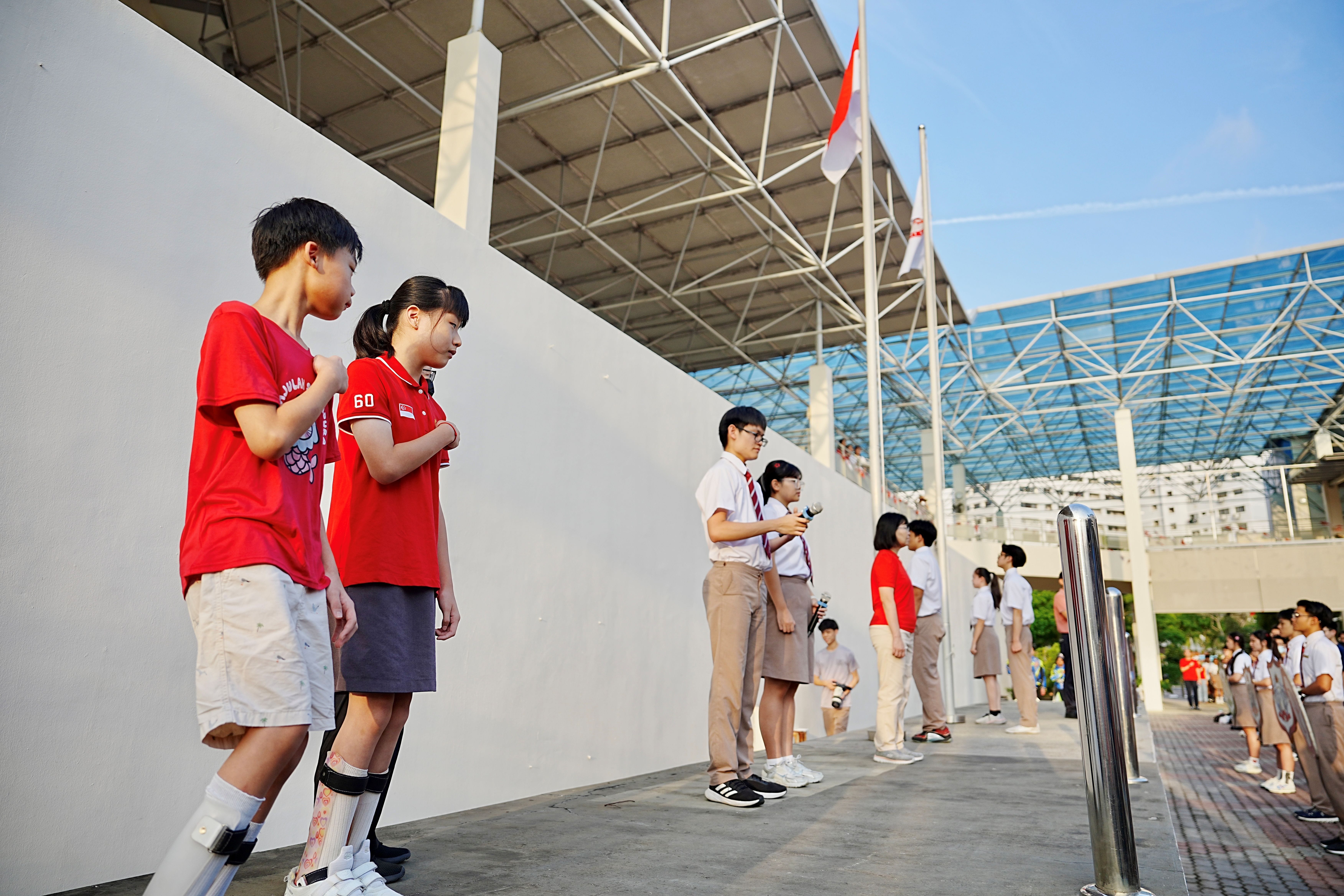 Children and students stand solemnly with fists to their chests, taking the National Pledge beneath a raised Singapore flag.